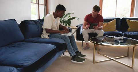 Two men collaborating on laptop and tablet on navy sofa in bright modern living room