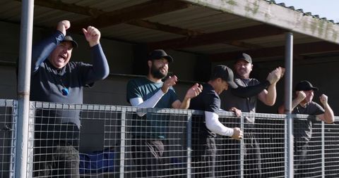 Male sports supporters cheering behind mesh fence at covered dugout showing team spirit