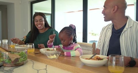 Smiling diverse family sharing mealtime in sunlit modern kitchen, child eating pasta