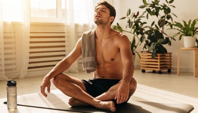 Shirtless man practicing breathwork on yoga mat after workout in sunlit home studio