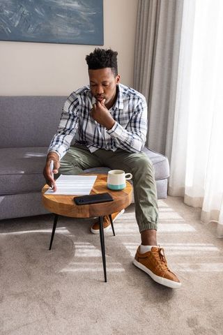 African american man contemplating paperwork at home pointing at document with coffee mug