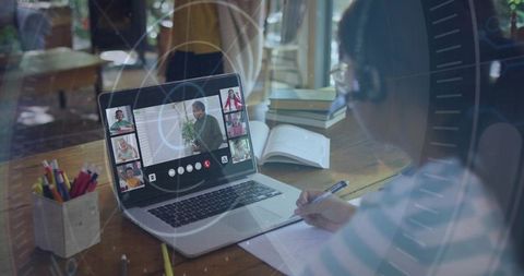 Child studying online with headset and laptop at home desk taking notes and learning remotely