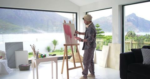 Indian artist painting at waterfront studio with large windows and mountain view