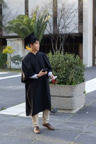 Graduating asian student wearing cap and gown holding diploma and smartphone on campus