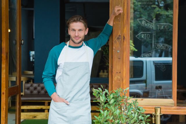 Portrait of smiling waiter standing with hand in pocket at entrance of cafe