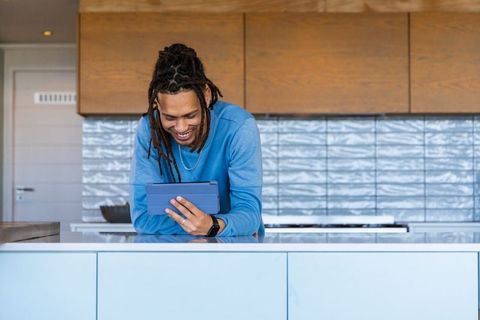 Smiling african american man leaning on white kitchen island using tablet and smartwatch