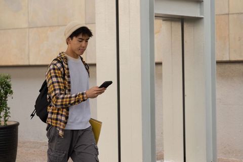 Asian student walking on campus using smartphone with backpack and yellow folder