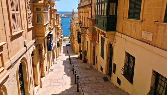 Sunlit maltese limestone stairway descending to blue harbor with traditional balconies