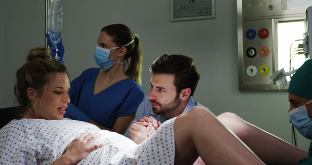 A Caucasian woman in labor is comforted by a man, presumably the father, as a nurse and a healthcare professional assist during the delivery, with copy space. The setting suggests a hospital delivery room, capturing a moment of support and anticipation during childbirth.