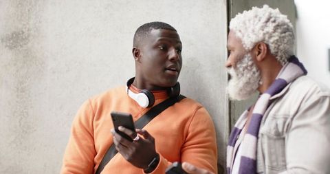 Young man sharing smartphone while chatting with older man by concrete wall