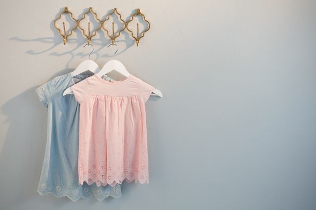 Two children's dresses, one blue and one pink, hanging on decorative hooks against a white wall. Ideal for use in articles or advertisements related to children's fashion, home organization, interior decor, or nursery room design.