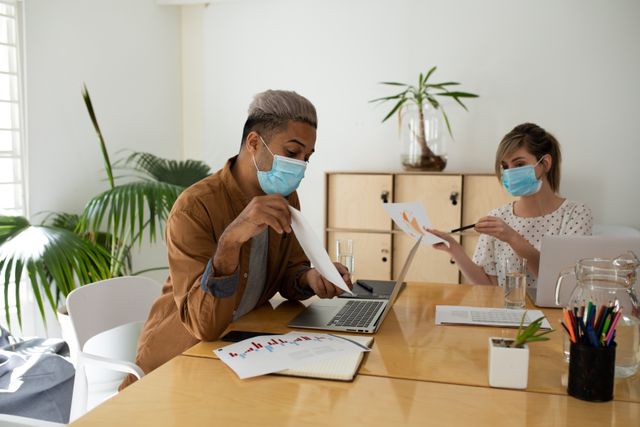 Caucasian man and woman having a meeting at an office while wearing facemasks. both of them are showing each other papers with graphs. on the table is a laptop, pens, and papers with graphs and stats.