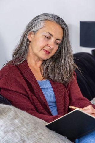 Mature woman journaling on sofa in cozy living room wearing maroon cardigan
