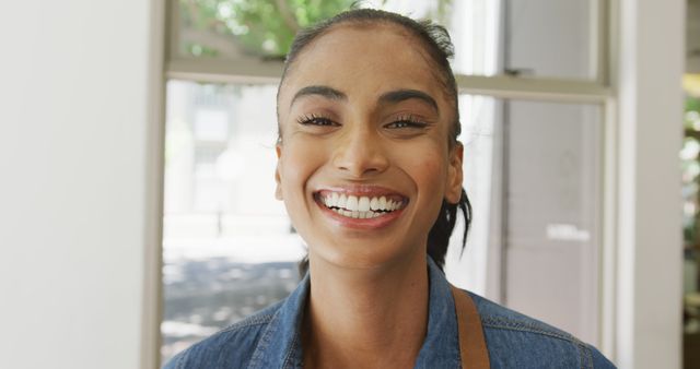 This image shows a cheerful young woman smiling brightly while wearing a casual denim shirt. Natural light illuminates her vibrant expression, giving a warm and friendly vibe. This image can be used for promoting positive messages, mental health awareness, casual fashion, or in professional contexts where approachable and confident personas are desired.