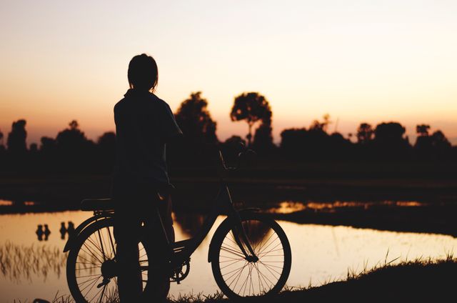 Person standing with bicycle by lake during sunset, offering a vibe of peace and contemplation. Ideal for themes of tranquility, outdoor lifestyle, rural life, solitude, mindfulness, nature appreciation, and serene landscapes. Can be used in advertisements, lifestyle blogs, or travel promotions.