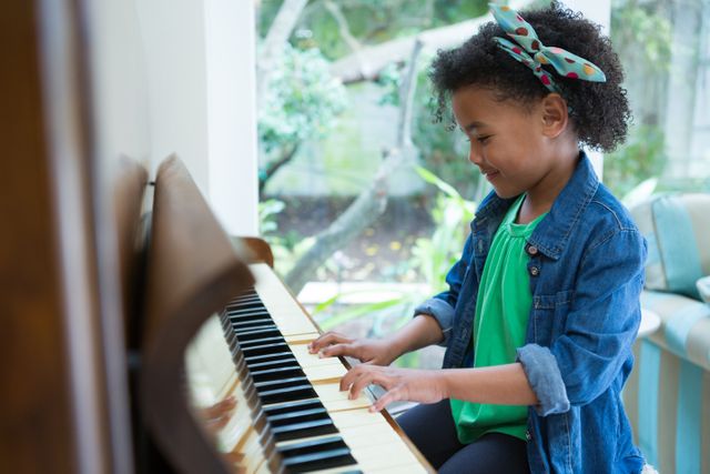 Young girl with curly hair playing piano at home, smiling and enjoying the music. She is wearing a green shirt and denim jacket, showcasing her talent and creativity. Perfect for educational materials, music lessons, and family-oriented content.