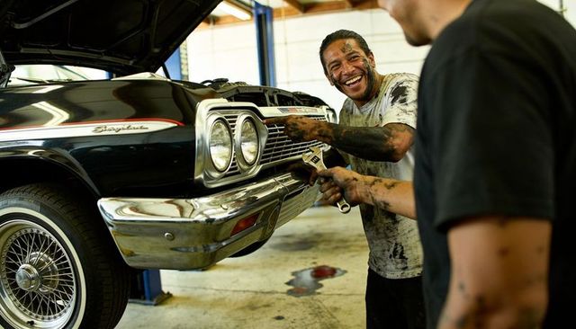 Grinning grease-stained mechanic holding wrench beside black vintage car with chrome bumper