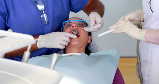 Dental professional examining patient's teeth with dental tools at clinic while assistant provides suction. Image useful for healthcare, dental hygiene, and oral health education. Suitable for dental clinics, medical brochures, or health-related articles.
