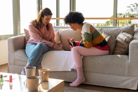 Multiracial women painting nails on sofa during cozy sunlit at-home manicure and chat