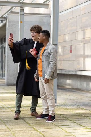 Graduating student taking selfie with friend on campus walkway holding diploma
