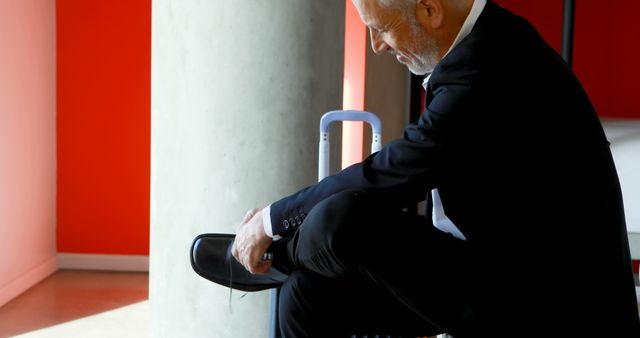 Senior businessman in formal suit tying shoes while sitting inside office building. He is preparing for work or business travel. Ideal for concepts related to professionalism, business routine, corporate environment, and senior executives in the workplace.