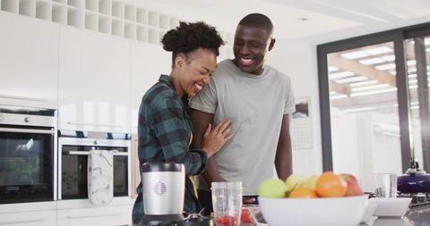 Laughing couple leaning close at kitchen counter with blender, strawberries and fruit bowl