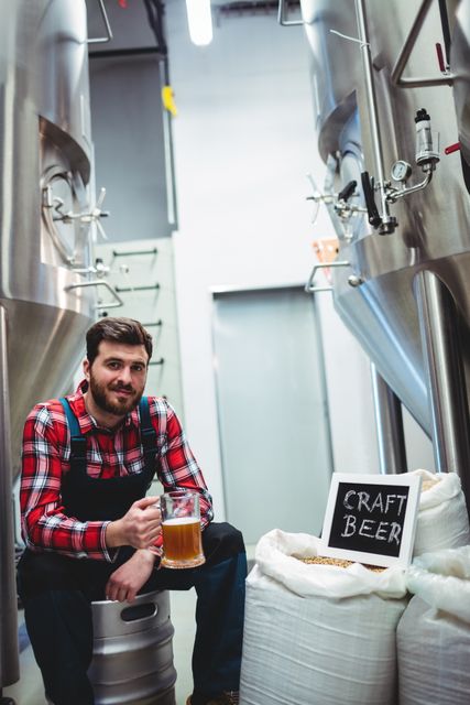 Male brewer in plaid shirt holding a glass of craft beer while sitting by storage tanks in a brewery. Ideal for use in articles about craft beer production, brewing processes, small breweries, and the beer industry. Perfect for marketing materials for breweries, brewing courses, and beer-related events.