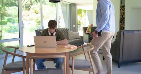 Teen working on laptop at wooden dining table in sunlit open-plan family living space