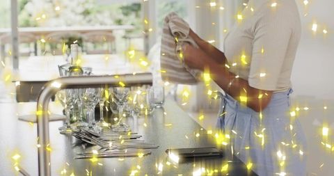 Woman drying wine glasses at sunlit kitchen counter wearing apron and holding towel