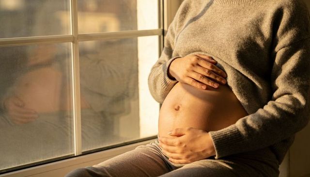 Pregnant woman cradling baby bump by sunlit window, cozy knit sweater, warm glow