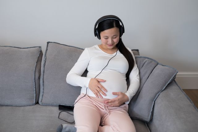 Pregnant woman sitting on a couch, wearing headphones, and listening to music. She is gently holding her baby bump and appears relaxed and content. This image can be used for articles or advertisements related to pregnancy, prenatal care, relaxation techniques for expecting mothers, or maternity products. It conveys a sense of calm and bonding between the mother and her unborn child.