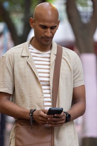 African man checking smartphone on urban plaza wearing striped shirt and messenger bag