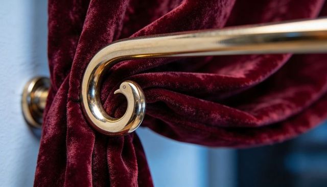Polished brass scroll holdback clasping burgundy velvet drapery, luxury interior detail