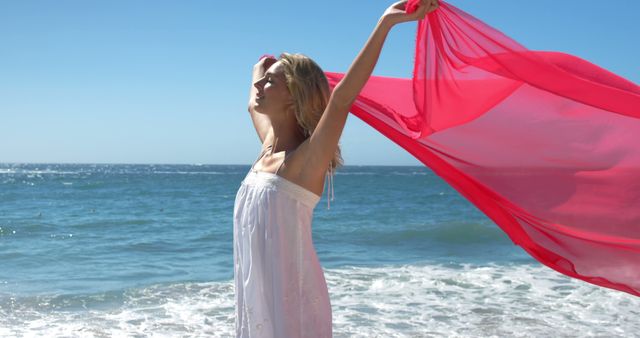 Blonde woman standing on sandy beach with ocean waves in background, holding flowing pink fabric as it sways in breeze. Perfect for illustrating concepts of relaxation, vacation, freedom, joy, and summer. Ideal for promoting travel destinations, wellness retreats, and summer-related advertisements.