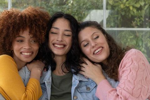 Three diverse friends hugging and laughing on couch wearing cozy sweaters by window