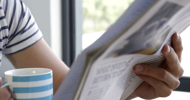 A person is casually reading a newspaper next to a window, with a coffee cup on the table, with copy space. Capturing a moment of relaxation, the image conveys a sense of leisure and staying informed.