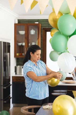 Mature woman decorating kitchen with green, gold and white balloons tying twine