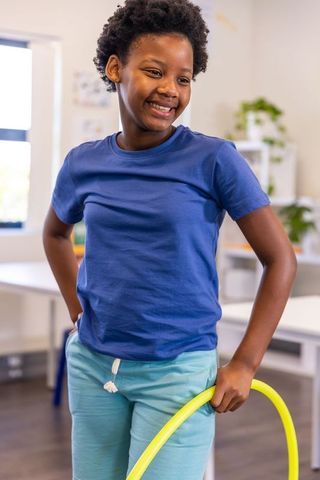 African american preteen girl smiling while holding yellow hula hoop in sunlit classroom