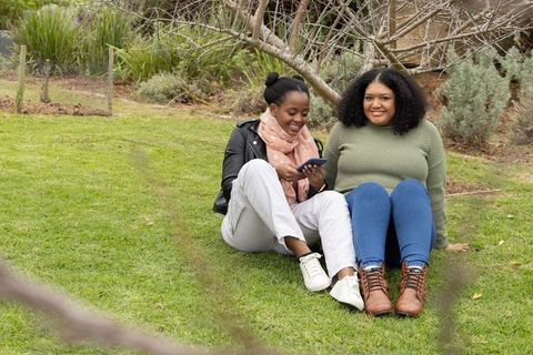 African american friends sharing smartphone while sitting on lawn in park casual autumn