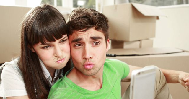 Young couple making silly faces and taking a selfie with cardboard boxes in the background. Picture captures fun and lighthearted moment during moving or unpacking in their new home. Great for use in real estate, moving services, or lifestyle blogs.