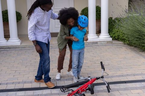 Black parents helping child learn bicycle riding after fall on paved suburban driveway