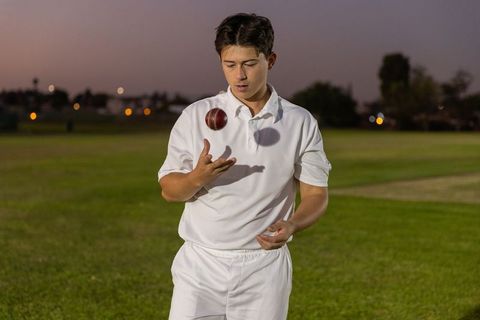 Cricket player tossing leather ball at dusk on grassy field in white kit
