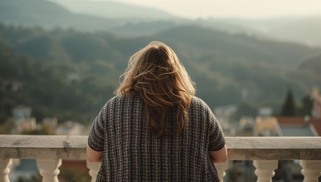 Woman gazing over valley from balcony wearing knit cardigan, rooftops and hills view