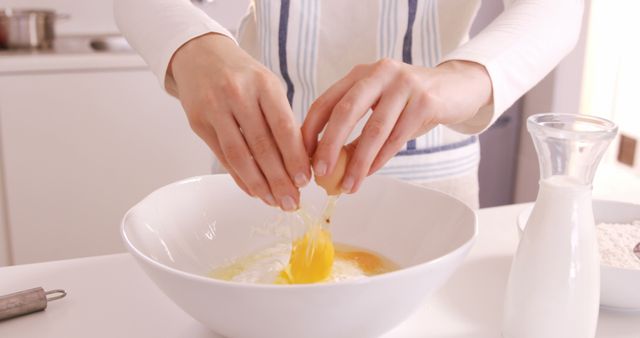 This image shows a person cracking an egg into a white bowl in a modern kitchen. Offering a close-up view, it captures the hands mid-action, emphasizing the baking process. Ideal for articles or advertising related to cooking classes, recipe blogs, culinary websites, or kitchen appliance promotions.