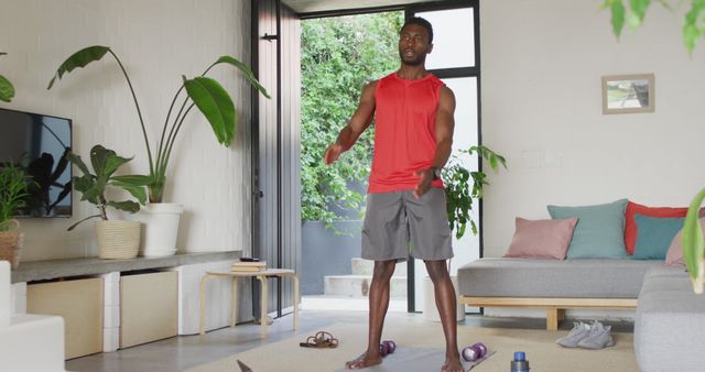 A man in a red tank top and shorts exercises in a modern living room using a laptop for online fitness instruction. The room features plants and minimalistic furniture, providing a comfortable home environment for a workout. This can be used in promotions related to home fitness, online fitness programs, healthy lifestyle motivation, or workout gear advertisements.