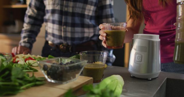 Couple seen making fresh green smoothies in modern kitchen. Vegetables, fruits, and blender shown on countertop, suggesting theme of healthy lifestyle and nutrition. Perfect for advertising kitchen appliances, healthy diets, or organic living.