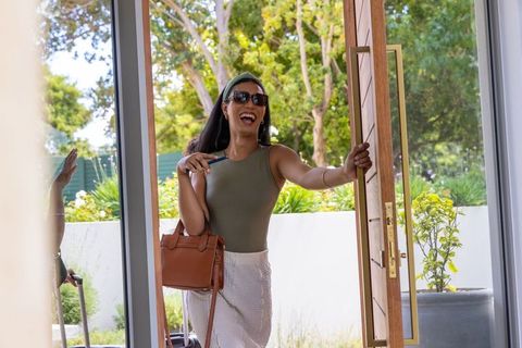 Laughing african american traveler entering modern home with suitcase and handbag