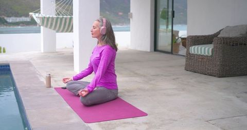 Mature woman meditating poolside on magenta mat wearing pink headphones and yoga clothes