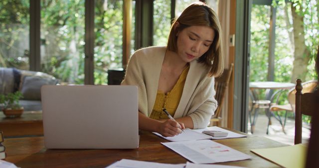 Woman is working on a laptop while writing on documents at a wooden table. Background shows a green, outdoor setting through large windows. Ideal for themes like remote work, productivity, home office lifestyle and casual work environment.