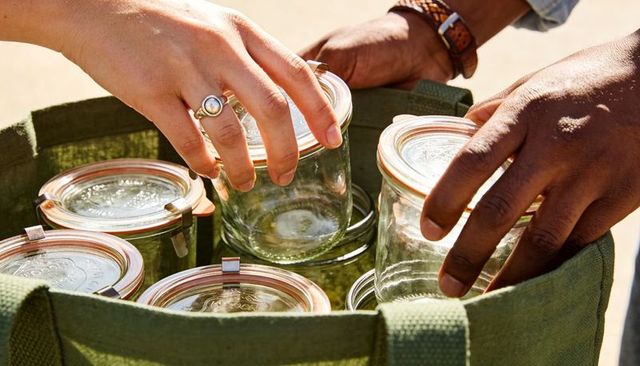 Two adults packing reusable canning jars into green canvas tote on sunlit porch